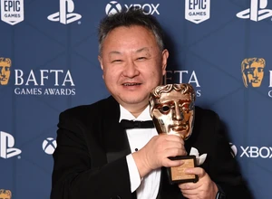 Мужчина в смокинге держит награду BAFTA на фоне синей стены с логотипами.