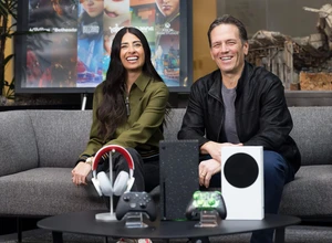 Два человека сидят на диване с игровой техникой и аксессуарами на столе.