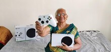 Пожилая женщина в сари держит игровой контроллер и консоль Xbox на диване.