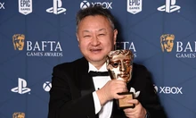 Мужчина в смокинге держит награду BAFTA на фоне синей стены с логотипами.