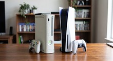 На столе стоят консоли Xbox 360 и PlayStation 5 рядом с геймпадами.