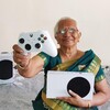 Пожилая женщина в сари держит игровой контроллер и консоль Xbox на диване.